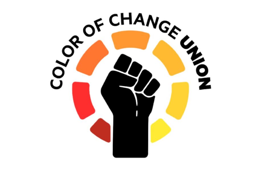 The image shows a stylized logo with the words "Color of Change Union" in black, forming an arc over a raised fist in black. The raised fist, a common symbol of solidarity and resistance, is surrounded by a half-circle of colored segments, ranging from red to yellow, symbolizing a gradient of change or diversity. The color gradient gives the design a dynamic feel, emphasizing empowerment and unity.